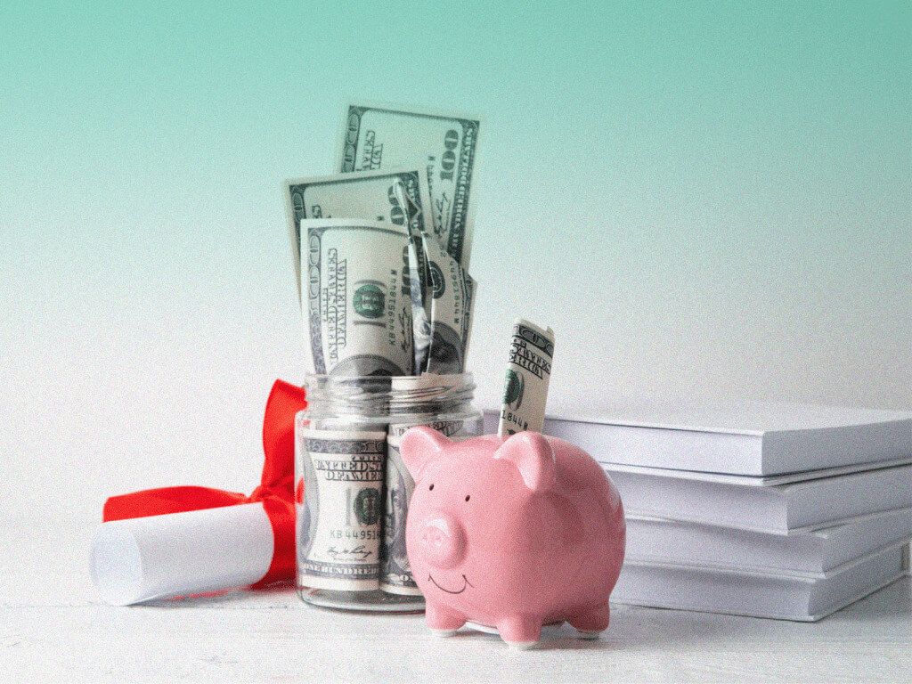 Cofre em formato de porquinho na cor rosa, ao lado de um pote de vidro aberto com notas de dinheiro dentro, um diploma de formatura enrolado com um laço vermelho em volta e alguns blocos de folhas de papel.
