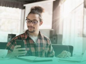 Estudante sentado à mesa olhando para o celular que segura em uma das mãos, utilizando um dos apps para estudar. Sua outra mão está sobre o notebook aberto e tem um caderno com um lápis em cima dele à frente do jovem.