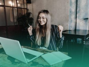 Estudante sentada à mesa sorrindo e comemorando uma boa organização de formatura com o site de formatura no notebook aberto à sua frente.