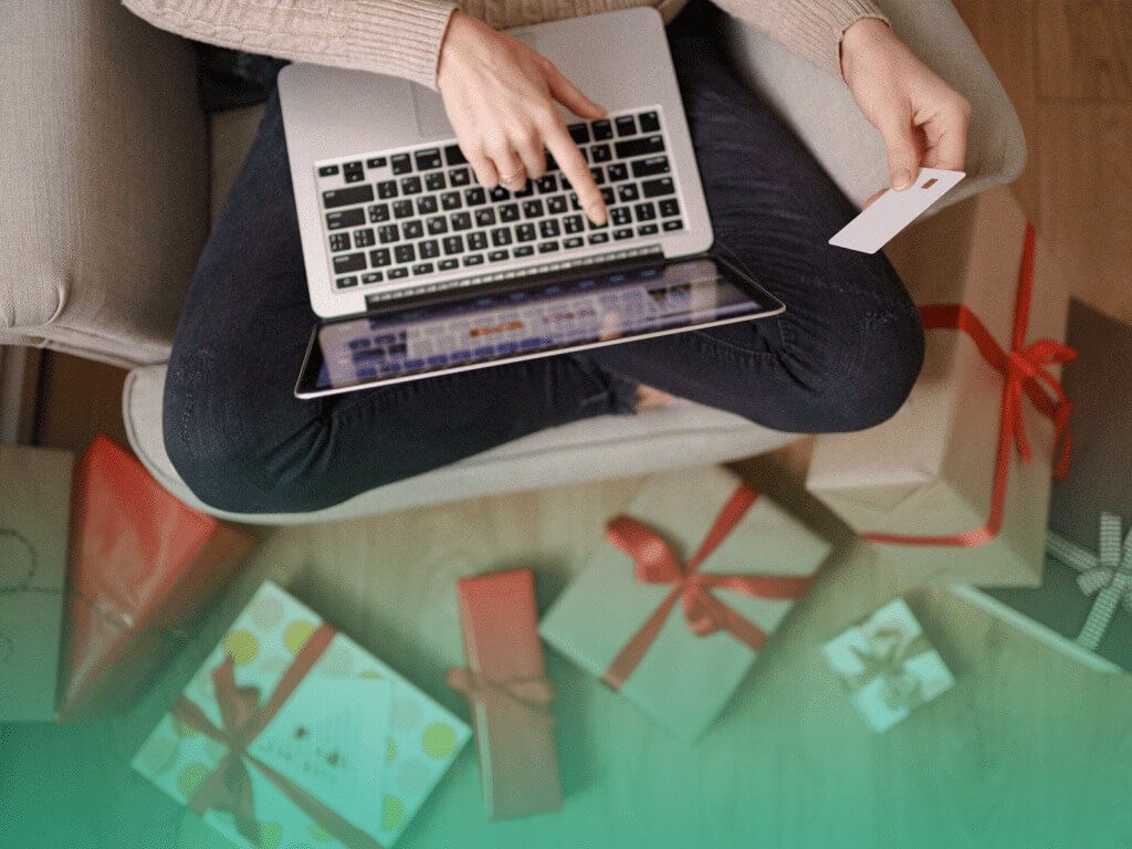 Mulher sentada de pernas cruzadas em uma poltrona, com um laptop no colo e um cartão de crédito em uma das mãos, comprando itens de uma lista de presentes virtual. No chão estão espalhadas algumas caixas de presente.