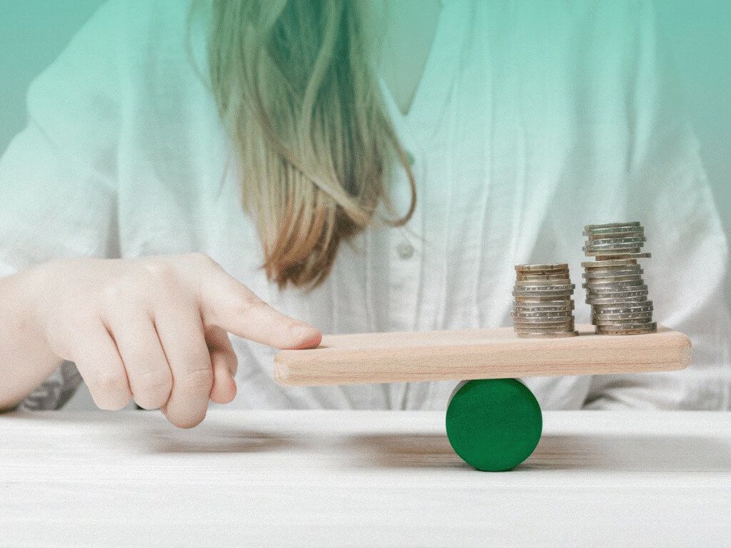 Mulher equilibrando, com o dedo indicador, algumas moedas em cima de um retângulo de madeira, que está apoiado sobre um objeto redondo e verde.