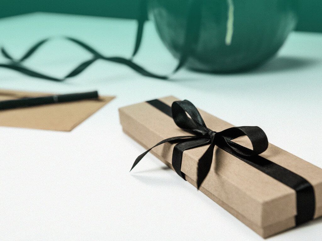 Uma caixa de presente fechada e amarrada com um laço preto sobre uma mesa. Ao fundo, um cartão em branco com uma caneta também preta sobre ele.