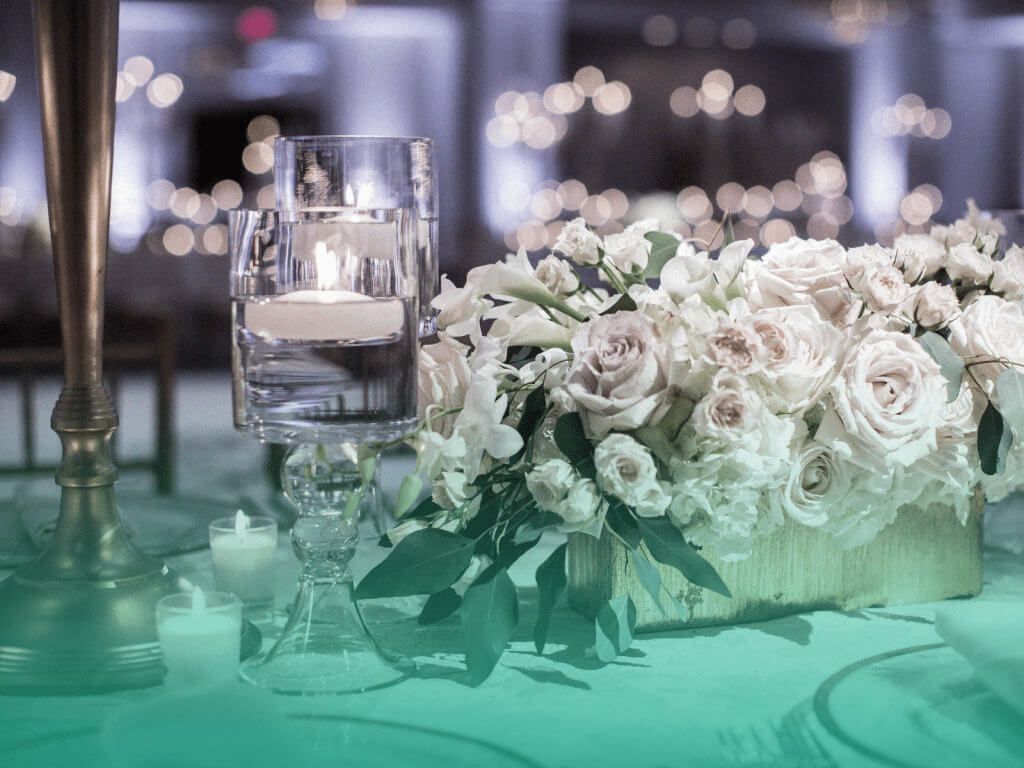 Recepção de formatura em ambiente interno, com foco nos enfeites de uma das mesas, com um arranjo de flores ao lado de um suporte alto de vidro com uma vela acesa dentro, sobre uma camada de água.