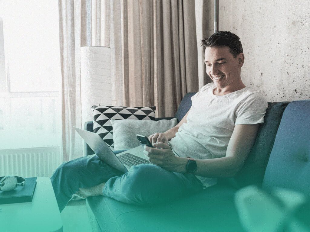 Jovem branco sentado em um sofá azul marinho com um laptop no colo e sorrindo para sua lista de presentes online na tela do celular que está segurando.
