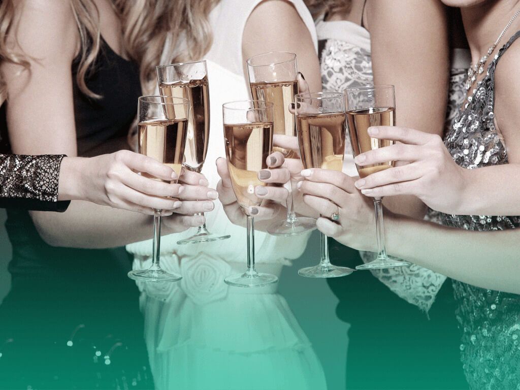 Seis mulheres em vestidos de festa segurando taças com bebida na frente do corpo, como se estivessem brindando. Os rostos das mulheres não aparecem.