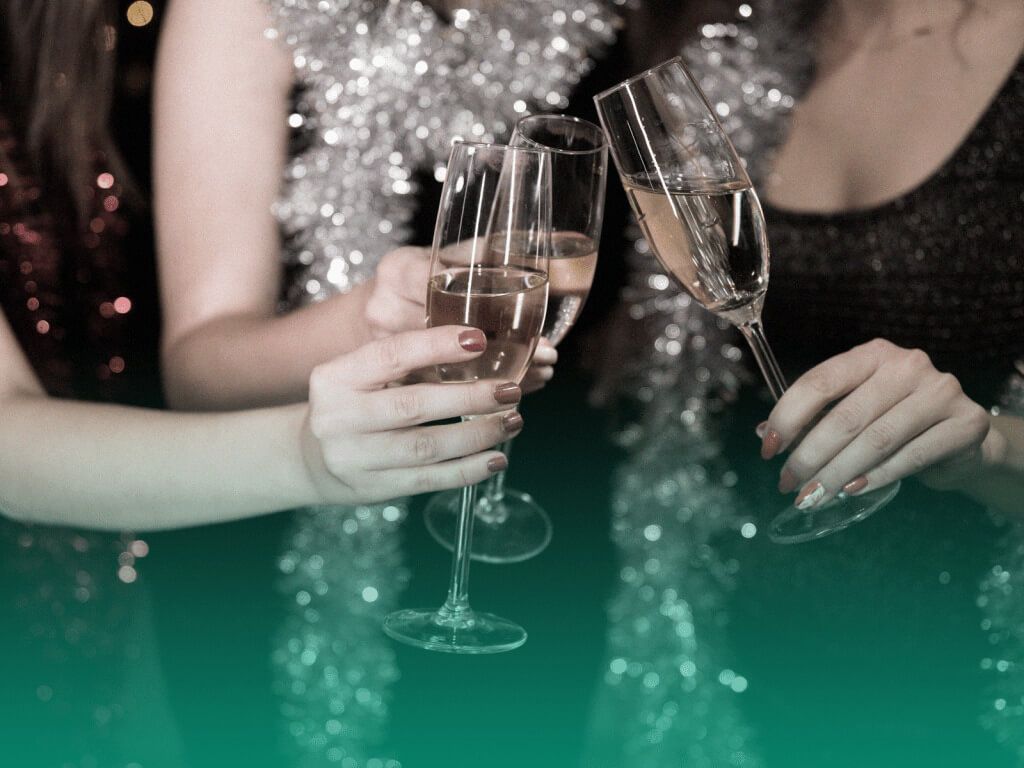 Três amigas com vestidos de festa em uma festa de formatura individual, juntando suas taças com bebida na frente do corpo em um brinde.