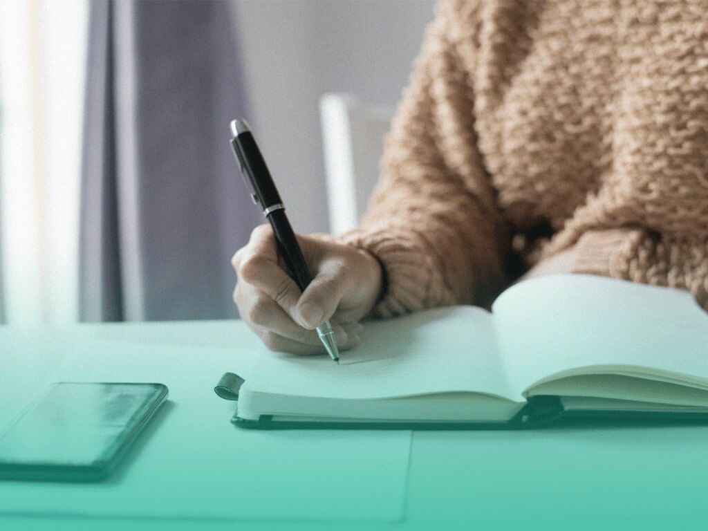 Foco na mão de uma mulher sentada à mesa escrevendo em um caderno, antes de descobrir sobre os benefícios da lista de presentes online.