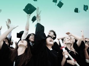 Grupo de estudantes diversos usando beca, sorrindo e jogando seus capelos para cima, comemorando a conquista de formatura.