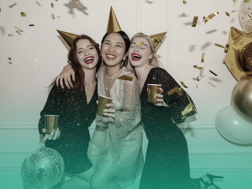Mulheres com vestidos de festa, sorrindo abraçadas com copos de bebida nas mãos e usando chapéus de festa, em um fundo branco com balões e confetes dourados, em uma recepção de formatura.