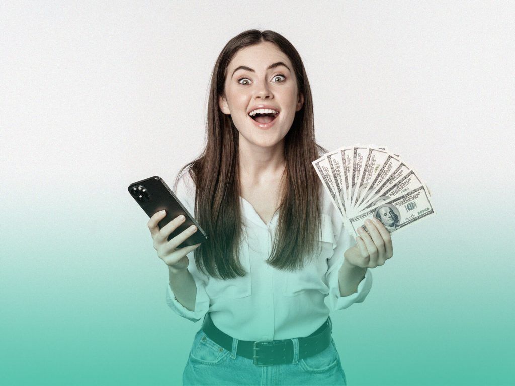Jovem de pé olhando para a câmera com uma expressão feliz, enquanto segura um celular em uma das mãos e um leque de notas de dinheiro na outra.