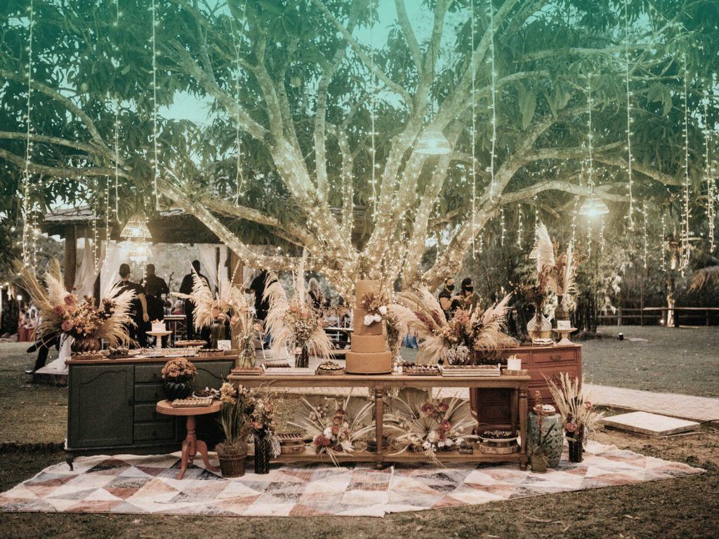 Decoração para formatura com fios com luzes pendurados em uma árvore, em uma festa externa, com convidados espalhados por uma extensa área verde atrás de uma mesa de madeira com um bolo de quatro andares decorado.