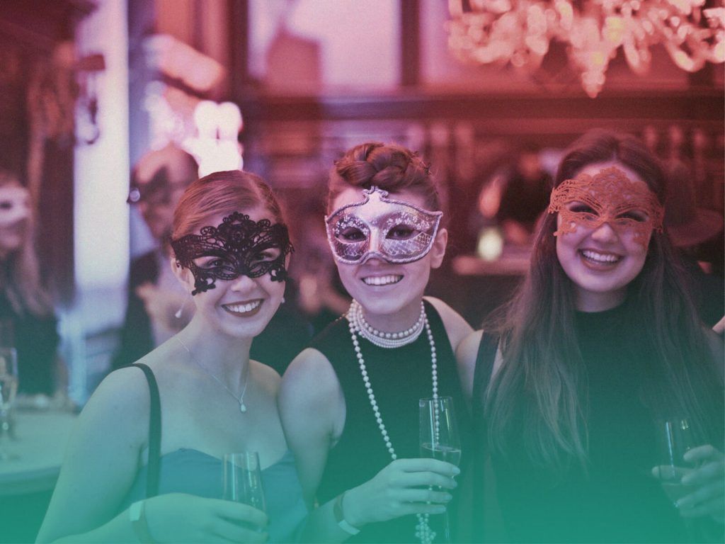 Três amigas usando máscaras, abraçadas de lado e sorrindo para a câmera com taças de bebida nas mãos, em uma festa com decoração para formatura temática. O ambiente está iluminado com luzes de cores quentes.
