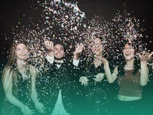 Amigos se divertindo juntos em uma festa de formatura, em um fundo preto e jogando confetes coloridos para cima.