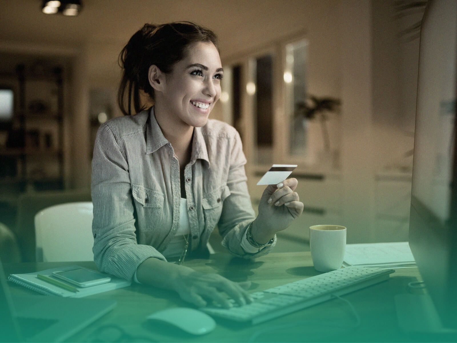 Mulher sentada à mesa e sorrindo enquanto olha para a tela de um computador, com uma das mãos sobre o teclado e a outra segurando um cartão de crédito.